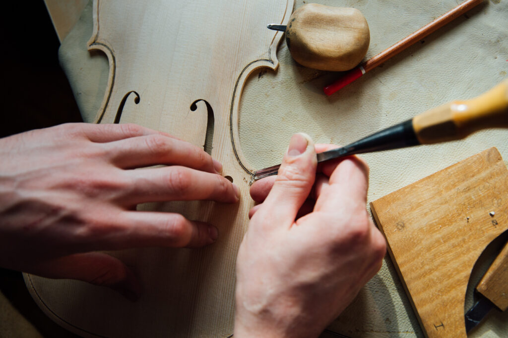 Master luthier's hands carving the f-hole of a violin — precision craftsmanship representing generational domain expertise that no AI model can replicate, the knowledge that makes MRO data classification accurate
