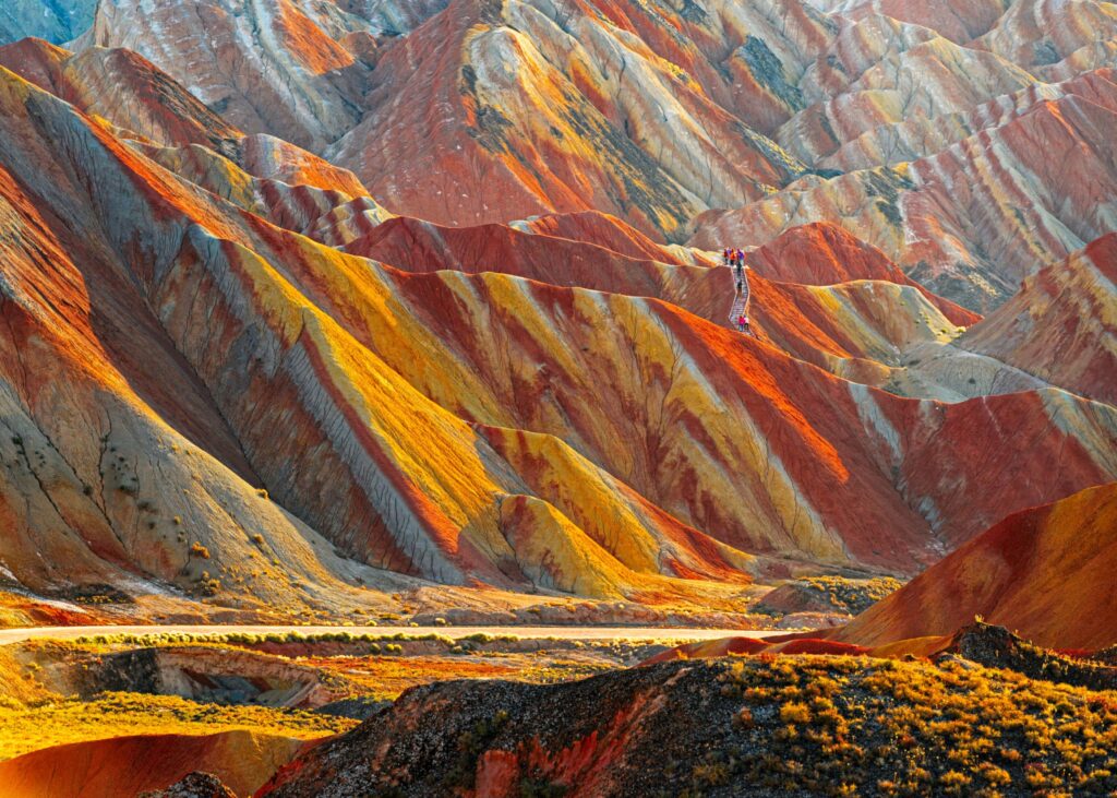 Vivid colour-banded mountain ridges at Zhangye Danxia, China — each geological layer distinct and visible, with a lone figure standing on the ridge providing scale against the vast layered landscape.
