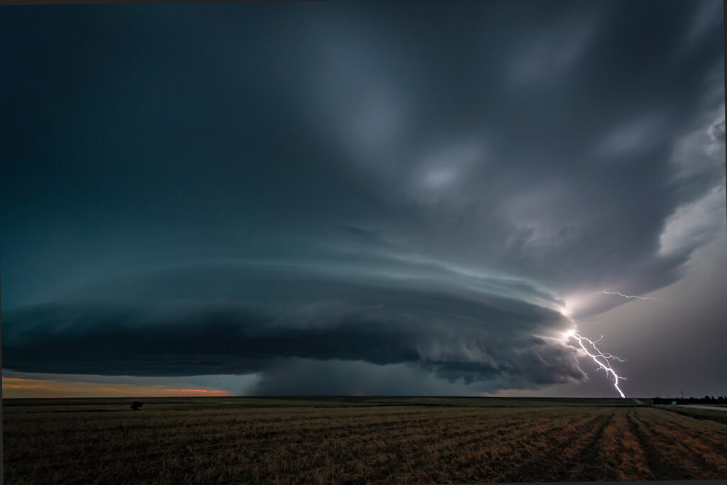 A lone cloud-to-ground lightning bolt striking empty plains under a dark sky, the circuit closing between an enormous charge and the earth — potential finally doing work once it finds its path to ground.